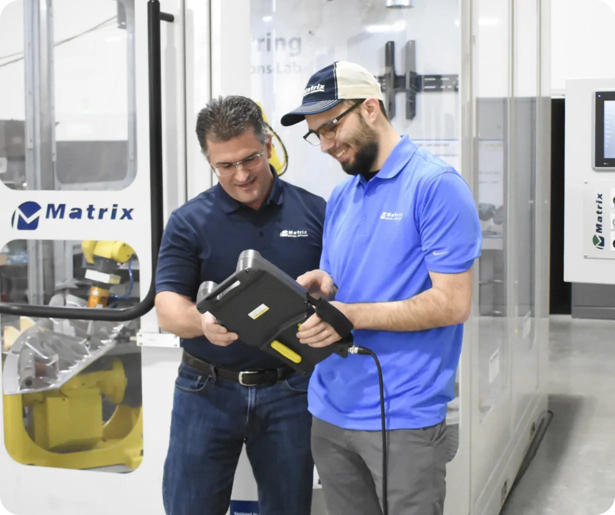 Two technicians in blue shirts review a rugged handheld tablet near Matrix machinery in a lab setting.
