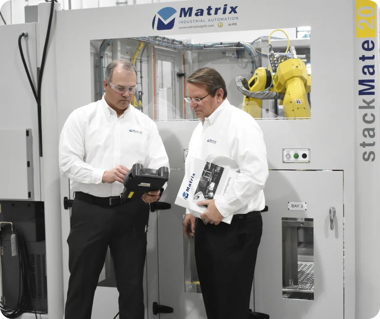 Two men in white shirts review a tablet and a brochure beside a large industrial machine labeled Matrix StackMat 20, in a factory setting.
