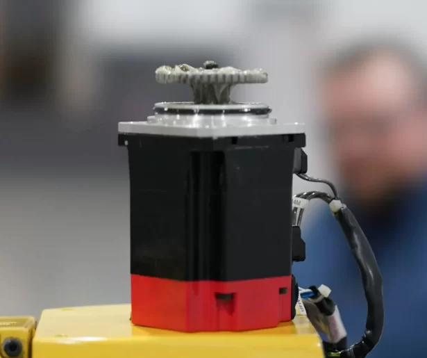 Close-up of a robotic actuator with a gear on top mounted on a yellow base; blurred workshop background.